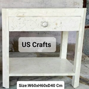 May include: White wooden nightstand with a single drawer and a lower shelf. The drawer has a floral design and a decorative knob. The nightstand measures 23.6 inches wide, 23.6 inches high, and 15.7 inches deep. The text "US Crafts" is visible on the front of the nightstand.