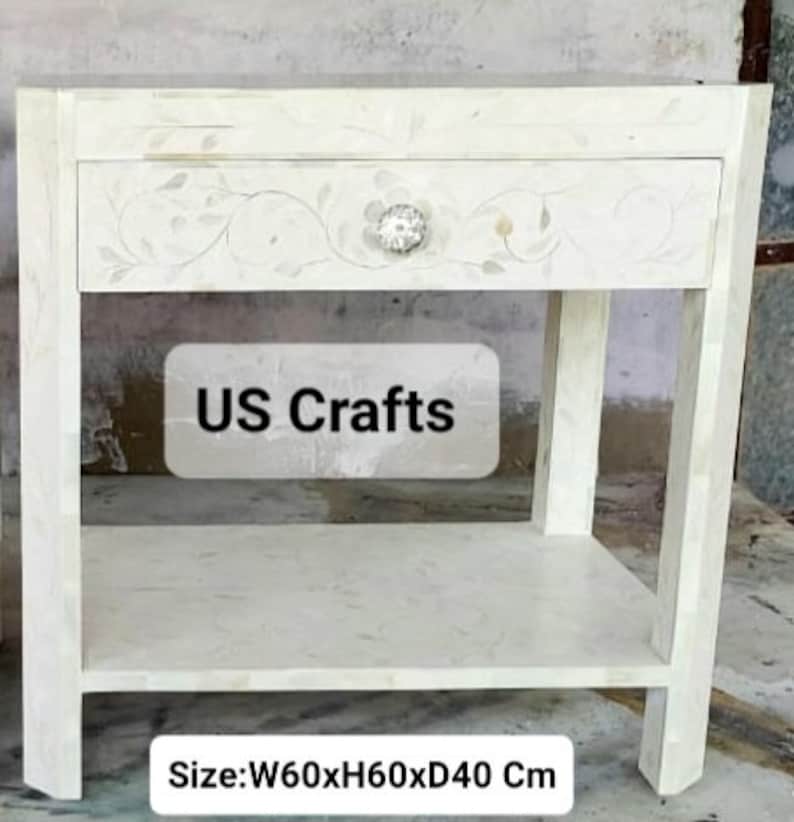 May include: White wooden nightstand with a single drawer and a lower shelf. The drawer has a floral design and a decorative knob. The nightstand measures 23.6 inches wide, 23.6 inches high, and 15.7 inches deep. The text "US Crafts" is visible on the front of the nightstand.