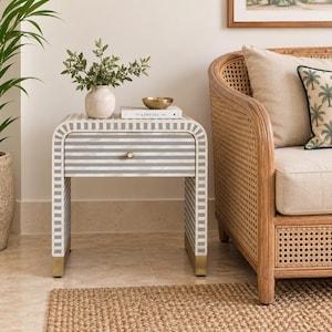 May include: A bedside table featuring a curved frame with a white and gray striped inlay pattern, a single drawer with a small circular knob, and metallic gold-toned feet. The table surface holds a small white vase with greenery and a book.