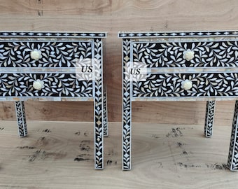 May include: Two black and white nightstands with intricate floral designs made of mother of pearl inlay. Each nightstand has two drawers with white knobs and stands on four legs.