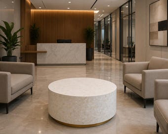 Op de afbeelding: Een ronde, lage salontafel met een wit, getextureerd oppervlak met een subtiel bladvormig patroon en een goudkleurige voet, geplaatst in een lobbyruimte met beige gestoffeerde fauteuils en een marmeren receptiebalie op de achtergrond.