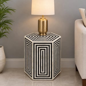 May include: A hexagonal side table featuring a black and white geometric striped pattern on its top and sides. A brass-toned table lamp with a hammered metal base and a cylindrical off-white fabric lampshade sits on the table surface.