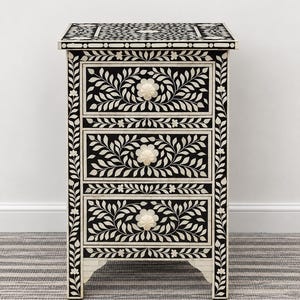 May include: A rectangular three-drawer chest featuring a black and white floral inlay pattern. The piece has a flat top surface, vertical side panels with leaf motifs, and three drawers, each centred with a round, off-white floral knob.