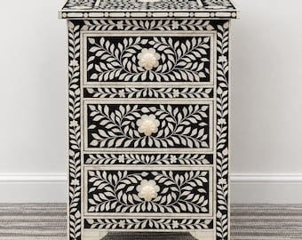 May include: A rectangular three-drawer chest featuring a black and white floral inlay pattern. The piece has a flat top surface, vertical side panels with leaf motifs, and three drawers, each centered with a round, off-white floral knob.
