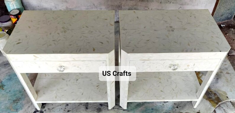 May include: Two white nightstands with a single drawer and a lower shelf. The nightstands are decorated with a floral pattern and have a crystal knob on the drawer. The text "US Crafts" is visible on the image.