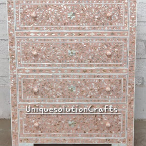 Puede incluir: Un aparador de madera rosa con cuatro cajones. Cada cajón está decorado con intrincados patrones florales hechos de incrustaciones de nácar. El aparador tiene un fondo blanco y está acentuado con ribetes dorados.