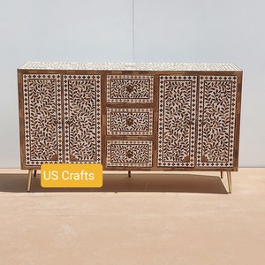 Può includere: Un mobile in legno bianco e marrone con intarsi floreali intricati e tre cassetti. Il mobile ha gambe in metallo dorato e il testo "US Crafts" è visibile sul davanti.