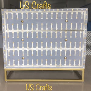 Puede incluir: Una cómoda de tres cajones con un acabado pintado en gris y un diseño de incrustaciones de hueso blanco. La cómoda tiene herrajes de color dorado y una base de metal dorado. La cómoda está etiquetada como "US Crafts".