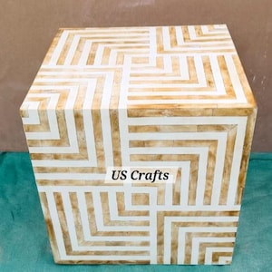 Può includere: Un tavolino quadrato con un motivo geometrico marrone chiaro e bianco. Il design presenta un motivo a zig-zag ripetuto sulla superficie. Il testo "US Crafts" è stampato sulla parte anteriore del tavolo.