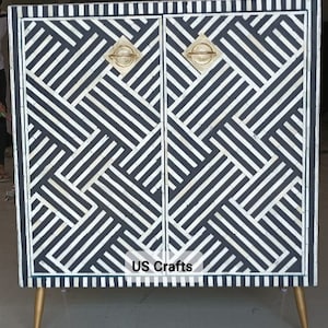 May include: A black and white geometric patterned cabinet with gold hardware and legs. The cabinet has two doors and the text "US Crafts" is printed on the front.