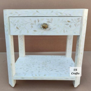 May include: White wooden nightstand with a single drawer and a lower shelf. The drawer has a floral design and a clear glass knob. The shelf also has a floral design. The nightstand is labeled "US Crafts".