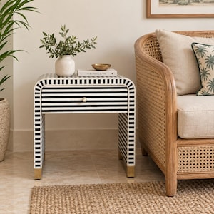 May include: A rectangular bedside table featuring a black and white striped pattern across its frame and single drawer. The table has a rounded top edge and is supported by four short, metallic gold-toned feet at the base of each leg.