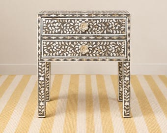 May include: A rectangular bedside table featuring two drawers with a dark gray finish and intricate white floral inlay patterns. Each drawer is equipped with a circular flower-shaped knob. The four straight legs also display matching floral inlay.