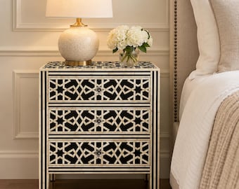 May include: A three-drawer nightstand featuring a black and white geometric star pattern with inlaid detailing. The piece has a rectangular frame with vertical striped legs and small, clear, round knobs on each drawer front for easy access.