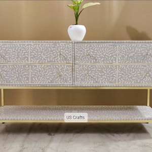 Puede incluir: Una cómoda blanca de incrustaciones de hueso con cuatro cajones y un estante inferior. La cómoda tiene un marco de metal dorado y un patrón floral en los cajones. La cómoda está etiquetada como "US Crafts".