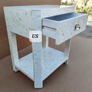 May include: White wooden nightstand with a single drawer and a lower shelf. The drawer has a floral design and a small, round, gold knob. The nightstand is decorated with a floral pattern.