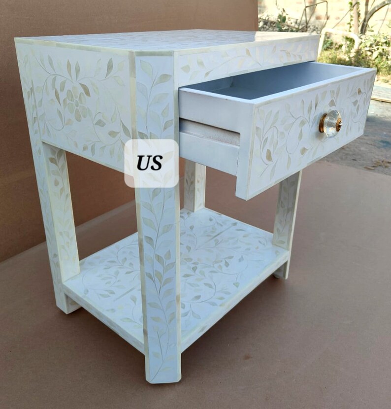 May include: White wooden nightstand with a single drawer and a lower shelf. The drawer has a floral design and a small, round, gold knob. The nightstand is decorated with a floral pattern.