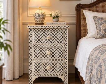May include: A rectangular bedside table featuring three drawers with a black and white geometric lattice pattern. Each drawer is equipped with a round, floral-shaped white knob. The table has a flat top surface and stands on four short legs.