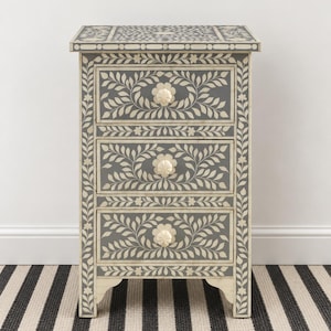 Puede incluir: Una mesita de noche de tres cajones con acabado gris oscuro e intrincados patrones de incrustaciones florales y de hojas en color crema. Cada cajón tiene un pomo redondo de color crema. El marco presenta un diseño de borde decorativo.