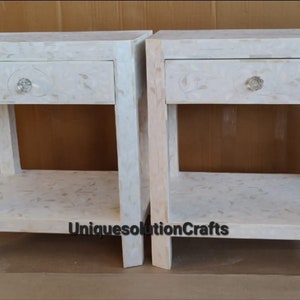 May include: Two white nightstands with a single drawer and a lower shelf. Each nightstand has a crystal-like knob on the drawer. The nightstands are made of a white material with a marbled pattern. The text "UniquesolutionCrafts" is visible on the bottom of the image.