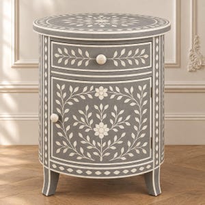 May include: A round gray bedside table featuring a single drawer and a cabinet door. The surface is decorated with intricate white floral and leaf patterns. It includes two small round knobs and stands on four curved legs with a matching inlay.