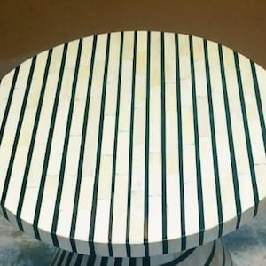 May include: Oval coffee table with a cream-colored surface and vertical black stripes. The table has a pedestal base with matching black stripes. A modern, decorative piece for a living room or lounge area.