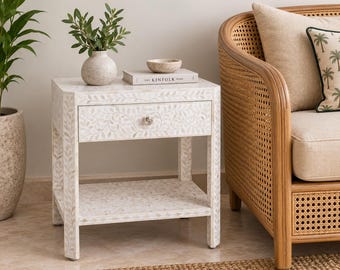 May include: A white bedside table featuring an intricate leaf pattern inlay, a single drawer with a round knob, and an open bottom shelf. The table is topped with a small ceramic vase containing green foliage and a book titled The Kinfolk Home.