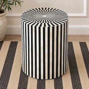 May include: A cylindrical side table featuring a high-contrast black and white striped pattern. The top surface displays a radial sunburst design of alternating black and white segments, while the sides are covered in vertical black and white stripes.