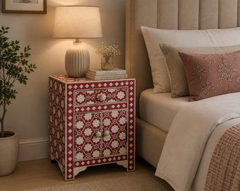 May include: A rectangular bedside table featuring a red and white geometric inlay pattern. The piece includes one top drawer and two lower cabinet doors, each fitted with small, round, light-colored knobs. The design displays a repeating star and diamond motif across the front and side panels, supported by a base with arched cutouts.