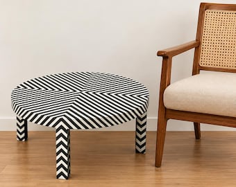 Op de afbeelding: Een ronde salontafel met een zwart-wit chevronpatroon op het blad en de poten. Daarnaast staat een houten fauteuil met een rugleuning van vlechtwerk en een lichtbeige gestoffeerde zitting, beide geplaatst op een houten vloer.
