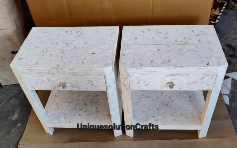May include: Two white nightstands with a floral design and a single drawer. Each nightstand has a lower shelf. The nightstands are made of wood and have a white finish.