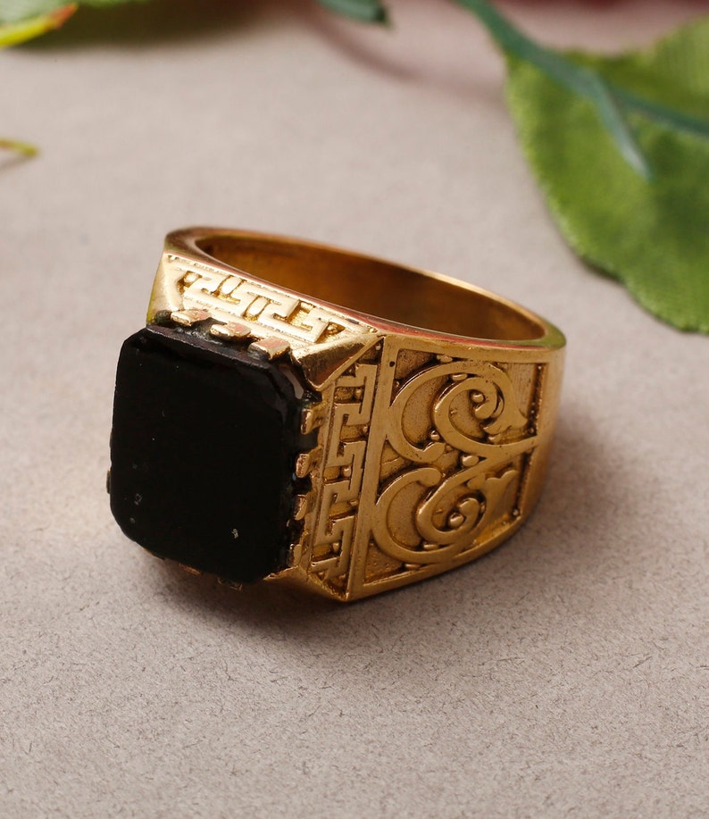 May include: A gold ring featuring a black stone and intricate detailing. The ring has a wide band with an ornate design.