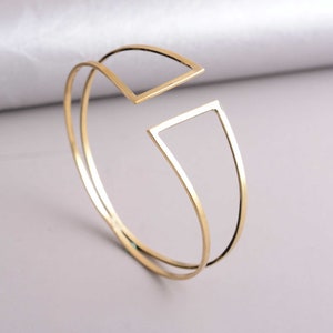 May include: A gold-toned double-layered bangle bracelet with a geometric design. The bracelet features a square shape with a cut-out section.