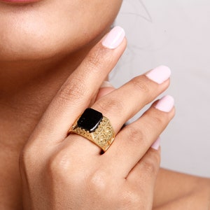 May include: A gold ring with a black square stone set in the centre. The ring has an intricate design around the stone.
