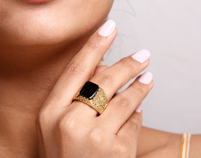 May include: A gold ring with a black square stone set in the centre. The ring has an intricate design around the stone.