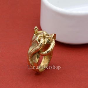 Könnte beinhalten: Ein goldfarbener Ring mit einem detaillierten Schlangendesign. Die Schlange ist um einen Schädel gewickelt, was ein einzigartiges und auffälliges Schmuckstück ergibt.