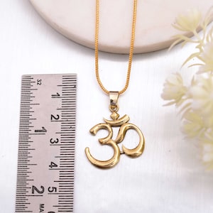 Puede incluir: Un collar con un colgante de símbolo Om de oro en una cadena de oro. El colgante mide aproximadamente 3,8 cm de alto y está colgado de una regla.