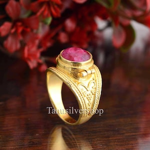 May include: A gold ring with a large, oval, pink gemstone. The ring has an ornate design on the band and the text "Tanusilvershop" is visible. The ring is set against a blurred background of red flowers.