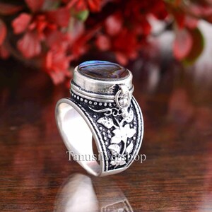 May include: A silver ring with a hinged compartment and a blue iridescent stone. The ring is decorated with a floral design.