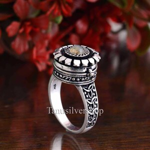 May include: A silver ring with a hinged compartment and a small, round, gray stone in the center. The ring is decorated with intricate designs and has a Celtic knot pattern on the band.