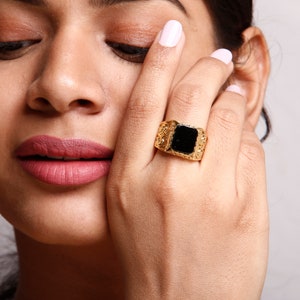 May include: A gold ring with a black square stone set in the centre. The ring has a textured band with intricate details.