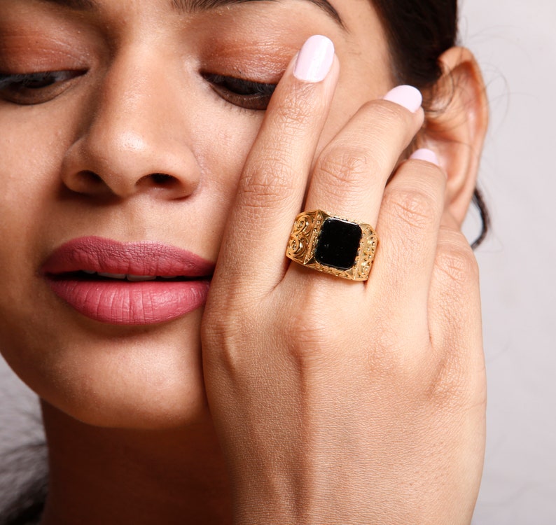 May include: A gold ring with a black square stone set in the centre. The ring has a textured band with intricate details.