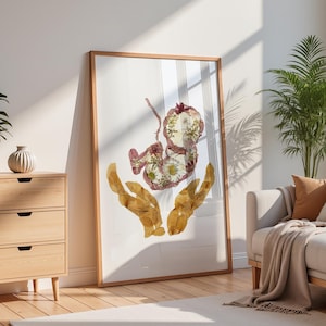 Könnte beinhalten: Ein gerahmter vertikaler Wandkunstdruck mit einer Collage aus gepressten Trockenblumen, die die Form eines Fötus in einer Gebärmutter bilden, gehalten von zwei Händen aus goldbraunen gepressten Blättern vor einem schlichten weißen Hintergrund.