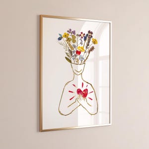 Può includere: Una stampa artistica da parete incorniciata con un disegno a linee dorate di un busto umano. La testa è sostituita da un colorato bouquet di fiori secchi pressati nei toni del giallo, blu e viola. Un cuore di fiore pressato rosso è sul petto.