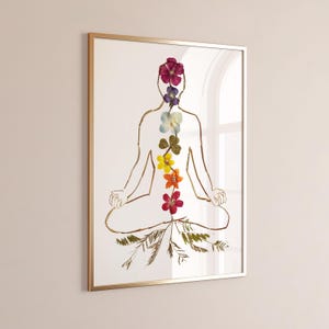 May include: A framed wall art print featuring a gold line drawing of a person in a seated meditation pose. The figure is filled with a vertical arrangement of seven pressed flowers in shades of purple, blue, green, yellow, orange, and red.