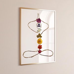 Può includere: Un'opera d'arte da parete incorniciata che raffigura una figura umana in una posa yoga seduta, realizzata con sottili ramoscelli marroni e fiori pressati. La figura include una composizione floreale viola, blu, azzurra, verde, gialla, arancione e rossa.
