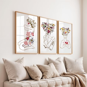 May include: Three framed wall art prints featuring gold line-art silhouettes of figures. The first shows a figure with pressed flowers on the body, the second a figure with a floral head, and the third a figure with a floral head and a red heart.