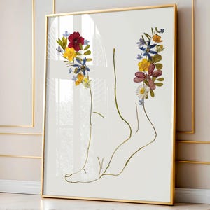Op de afbeelding: Een ingelijste wandkunstprint met een minimalistische gouden lijntekening van een menselijke voet en enkel. De omtrek is versierd met geperste gedroogde bloemen in tinten rood, geel, blauw en wit, geplaatst bij de tenen en het hielgebied.