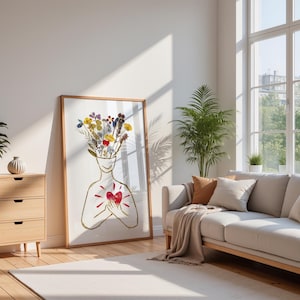 Könnte beinhalten: Ein gerahmter Kunstdruck mit einer goldfarbenen Strichzeichnung einer menschlichen Figur. Die Figur hat einen Strauß bunter Trockenblumen, der aus dem Kopf ragt, und hält eine leuchtend rote Herzform vor dem Brustbereich auf weißem Hintergrund.