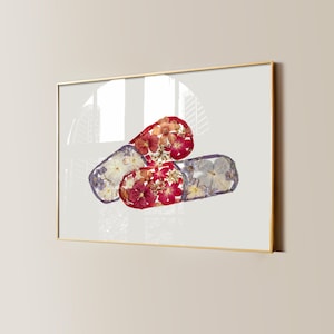 Op de afbeelding: Een ingelijst kunstwerk met drie abstracte vormen gevuld met geperste bloemen. De vormen zijn in tinten rood, wit en paars, met een gouden lijst. De achtergrond is lichtgrijs.